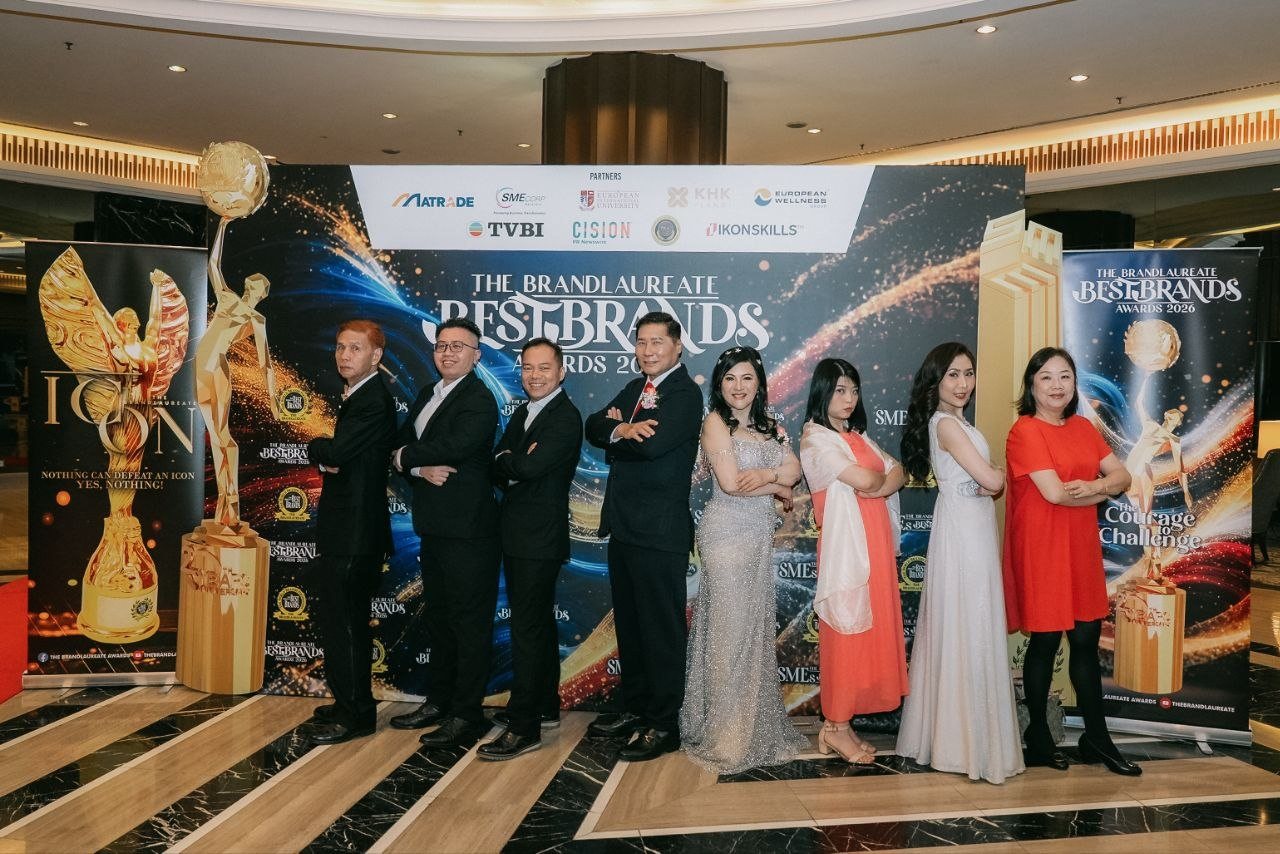 Left to right, three men of the team in black suits, DR. J AARON K DAVID in a navy suit and red tie, DR. SHEKINAIH SIEW SIN YAP in a silver beaded gown, a team member in a coral dress with a white shawl, Florence in an ivory beaded gown and a team member in a red dress pose with arms folded before The BrandLaureate BestBrands Awards 2026 backdrop.