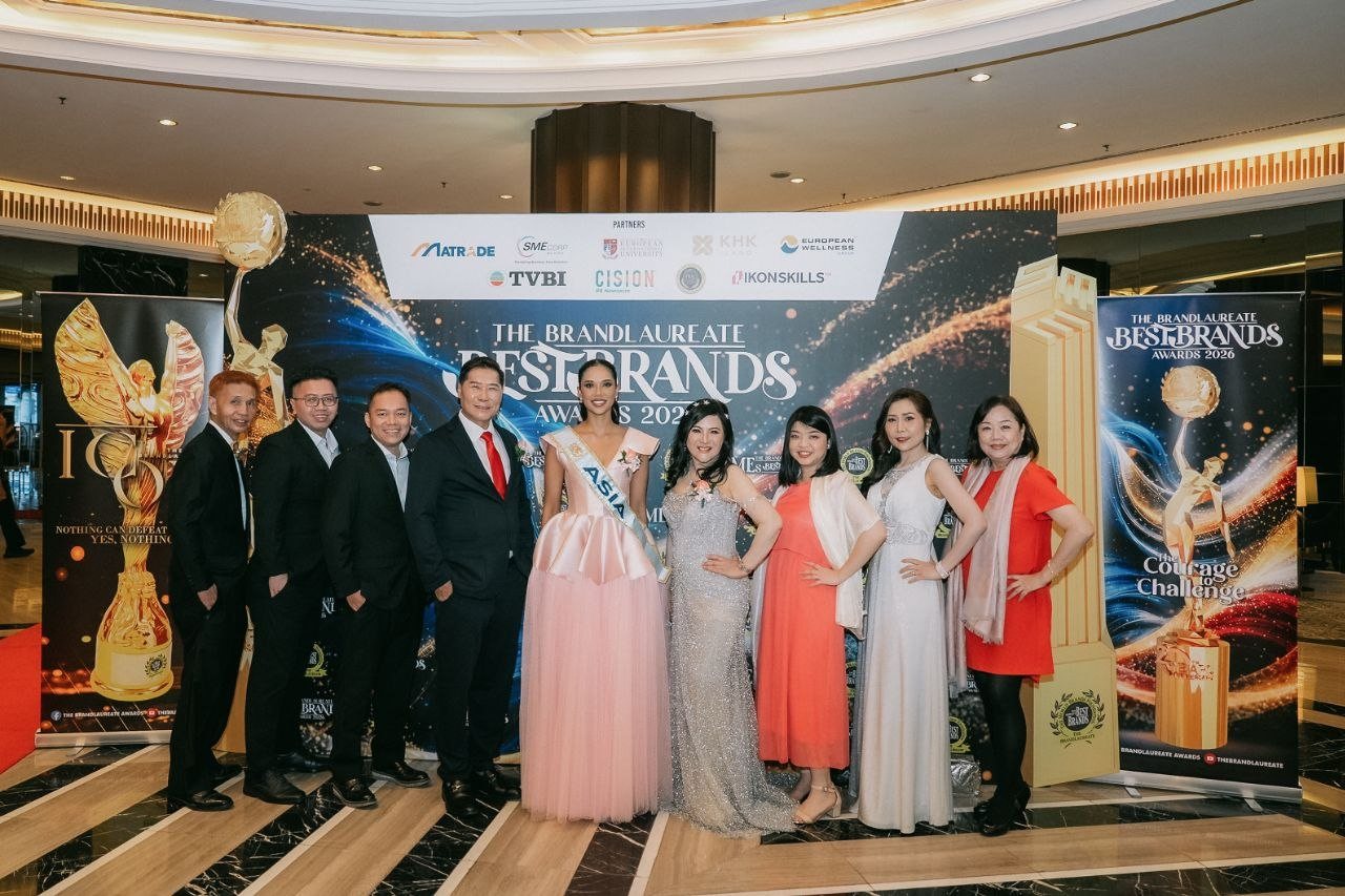 DR. J AARON K DAVID and DR. SHEKINAIH SIEW SIN YAP with the team of CE Global Network International and Crowned Eagles Global and Miss Universe Malaysia, in a pink gown and sash, before The BrandLaureate BestBrands Awards 2026 backdrop, between the gold trophy replicas