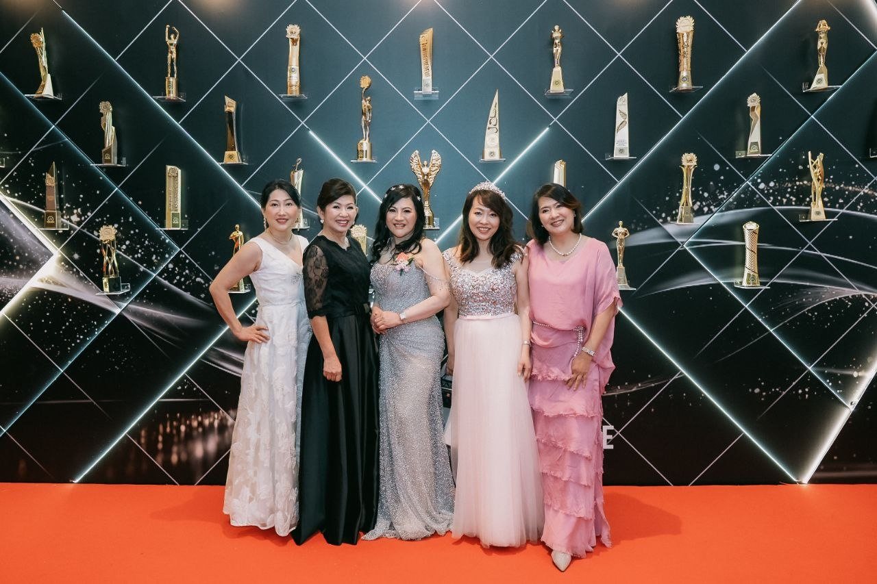 Left to right, a guest in a white lace gown, a key member of The BrandLaureate team in a black lace dress, DR. SHEKINAIH SIEW SIN YAP in a silver beaded gown, a guest in a pink tulle gown and tiara and a guest in a pink tiered dress stand on the red carpet before the illuminated awards wall of trophies.