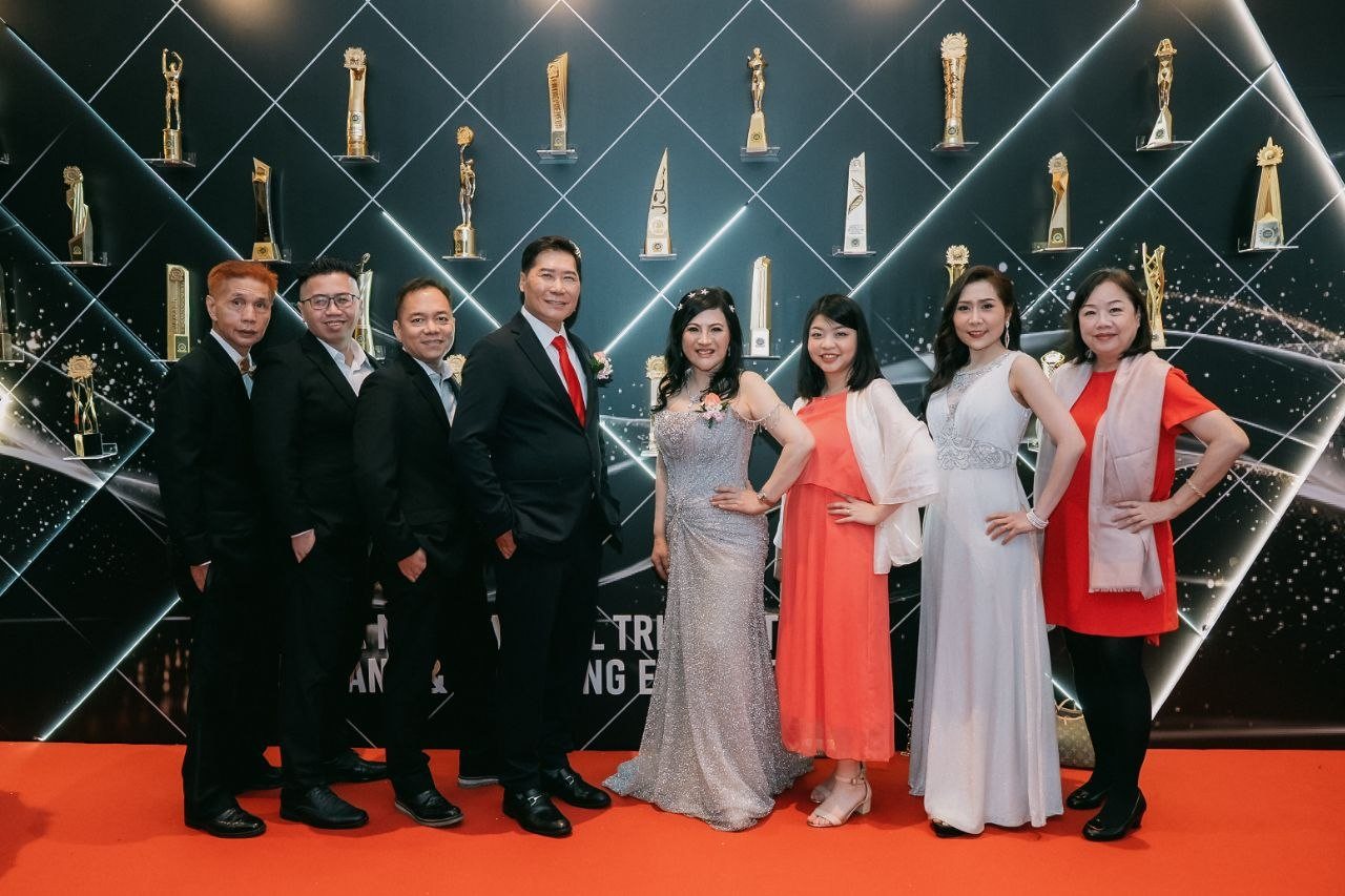 Left to right, three men of the team in black suits, DR. J AARON K DAVID in a navy suit and red tie, DR. SHEKINAIH SIEW SIN YAP in a silver beaded gown, a team member in a coral dress with a white shawl, Florence in an ivory beaded gown and a team member in a red dress with a pink shawl stand on the red carpet before the illuminated awards wall of trophies.