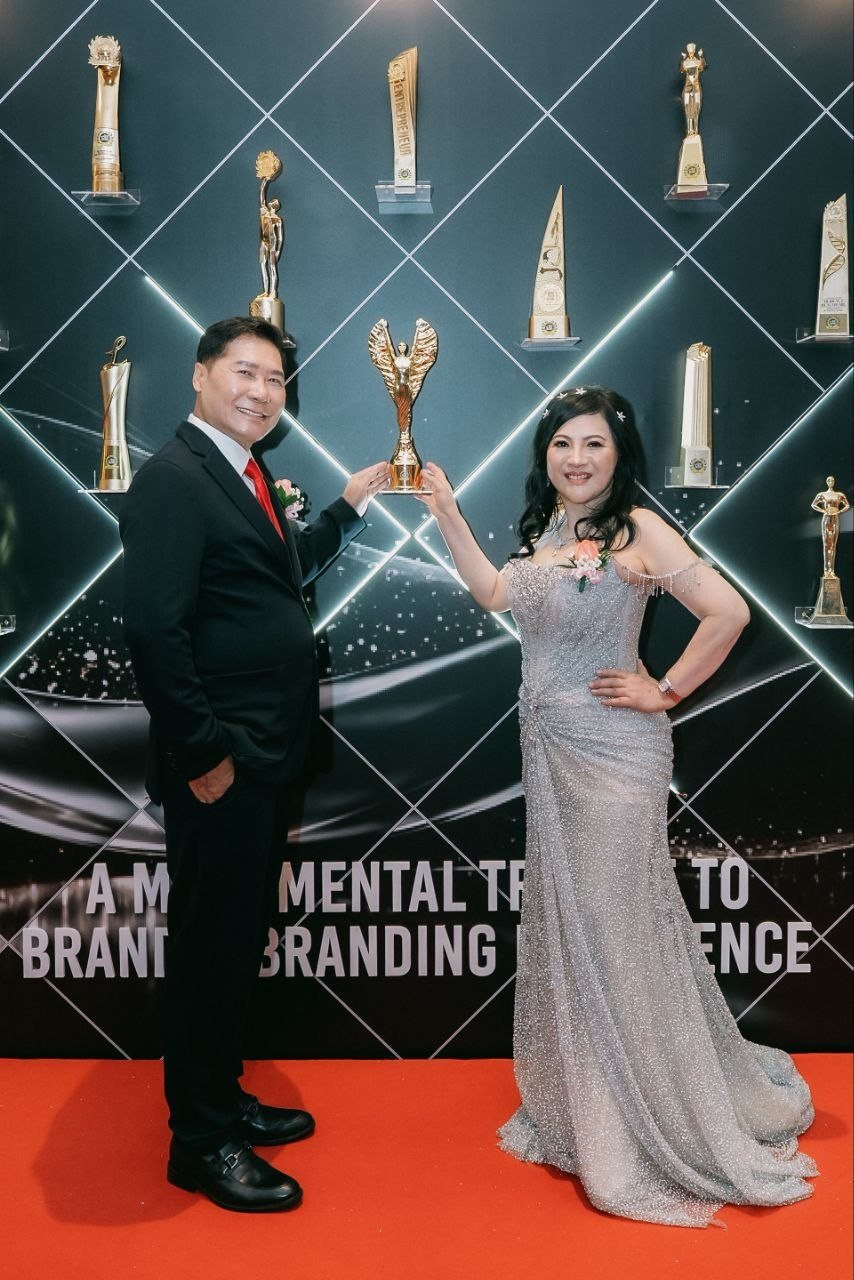 DR. J AARON K DAVID and DR. SHEKINAIH SIEW SIN YAP stand full-length on the red carpet before the awards wall of trophies, holding one gold winged trophy up between them