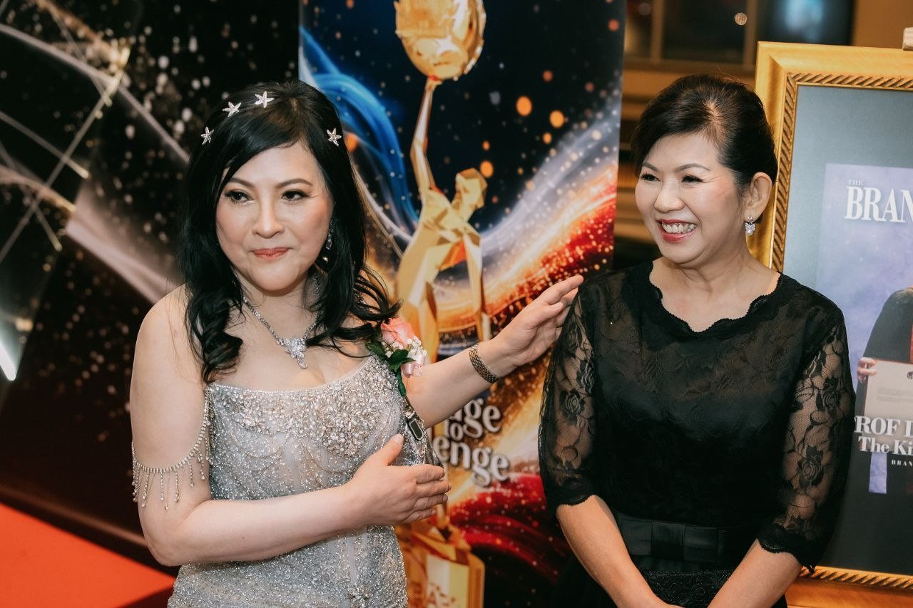 DR. SHEKINAIH SIEW SIN YAP, in a silver beaded gown with star pins in her hair and a pink corsage, rests a hand on the shoulder of a key member of The BrandLaureate team in a black lace dress, beside a BestBrands banner and a framed magazine cover.