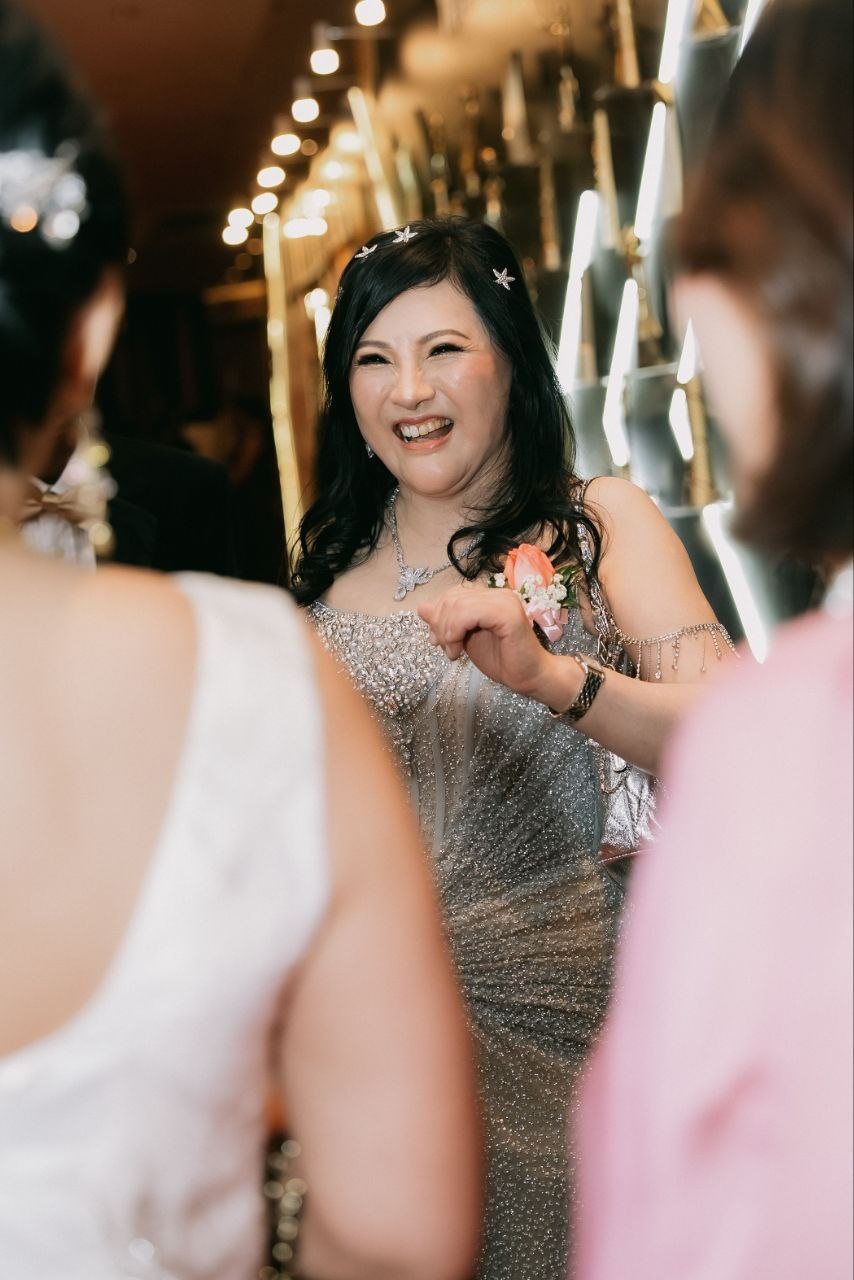 DR. SHEKINAIH SIEW SIN YAP, in a silver beaded gown with star pins in her hair and a pink corsage, laughs as she talks with guests, framed by two guests in the foreground before the lit awards wall.
