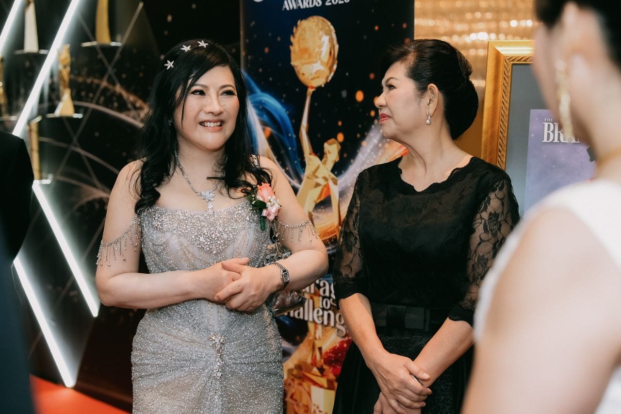 DR. SHEKINAIH SIEW SIN YAP, in a silver beaded gown with a pink corsage, smiles as she talks with a key member of The BrandLaureate team in a black lace dress, beside a BestBrands banner and a framed magazine cover.