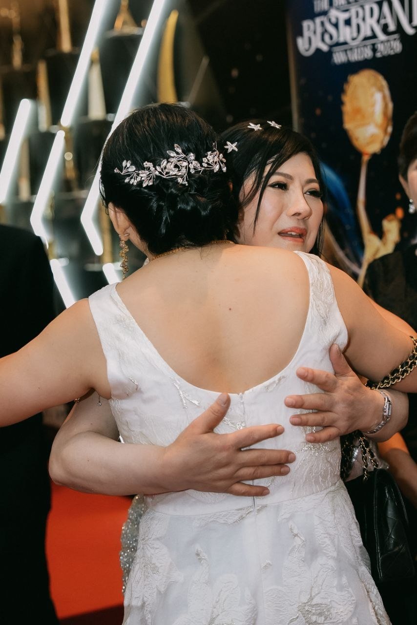 DR. SHEKINAIH SIEW SIN YAP, with silver star pins in her hair, embraces a guest in a white lace gown with a jewelled hair comb, before the lit awards wall and a BestBrands banner.