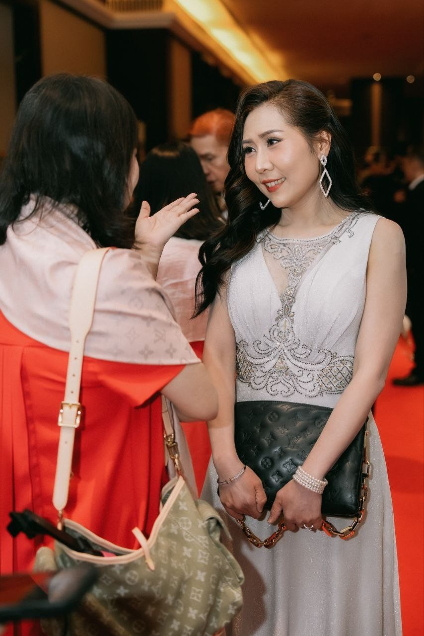 Florence, in an ivory sleeveless V-neck beaded gown holding a dark monogram clutch, smiles at a guest in a red dress and pink shawl on the red carpet.