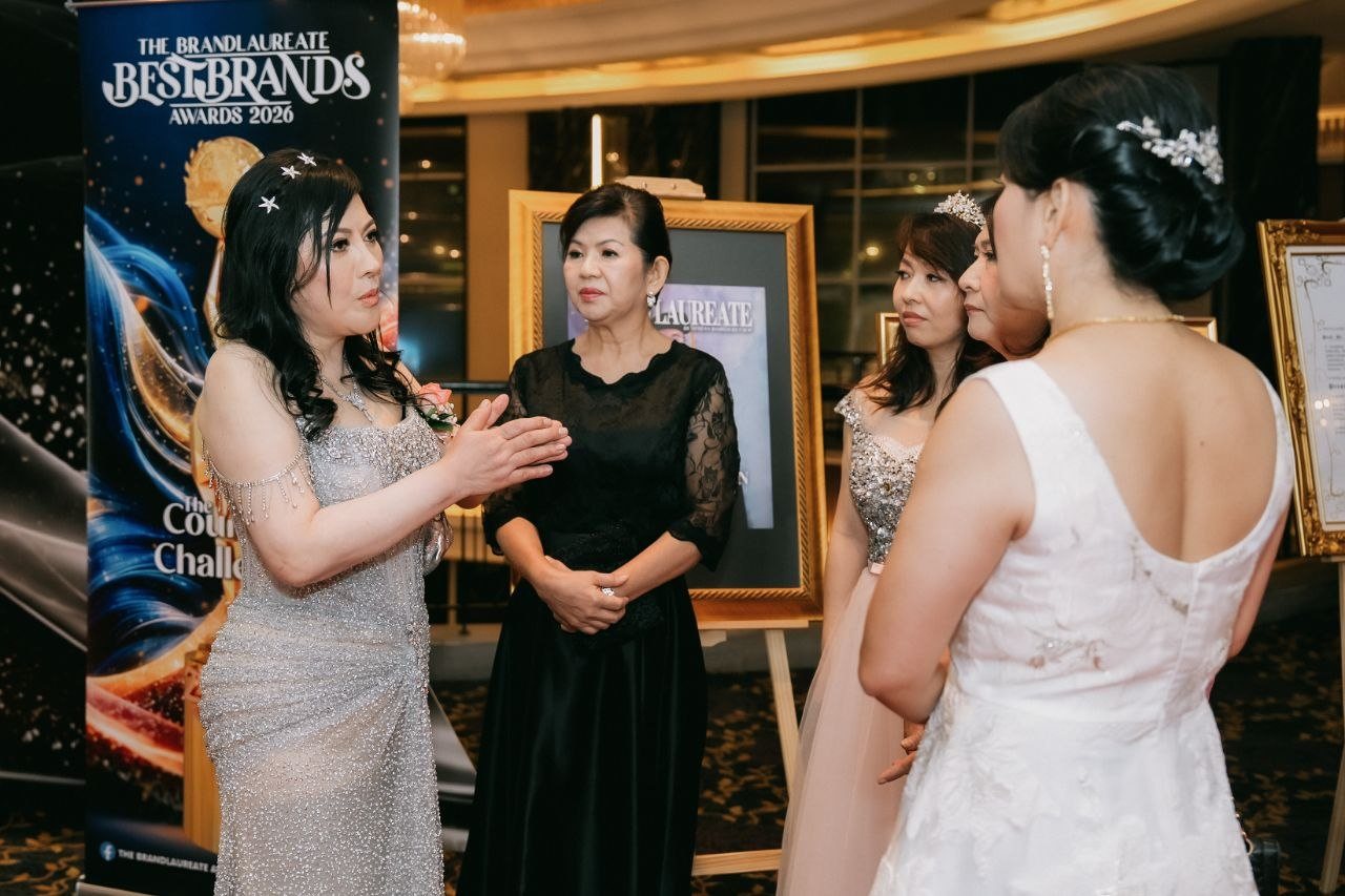 Left to right, DR. SHEKINAIH SIEW SIN YAP in a silver beaded gown gestures as she speaks to a key member of The BrandLaureate team in a black lace dress, a guest in a pink tulle gown and tiara, a guest partly hidden behind her, and a guest in a white lace gown seen from behind, beside a BestBrands banner and framed displays.