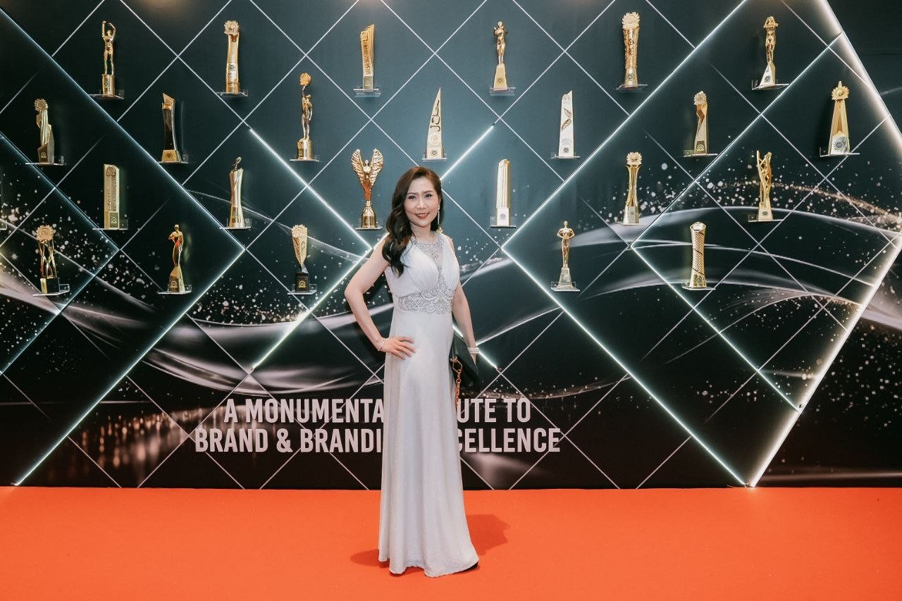 Florence, in a full-length ivory sleeveless V-neck beaded gown with a dark clutch bag, stands on the red carpet before the illuminated awards wall of trophies.