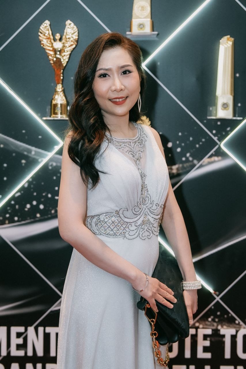 Florence, with long dark wavy hair and an ivory sleeveless beaded gown, smiles holding a dark clutch bag, with a gold winged trophy on the awards wall behind her.
