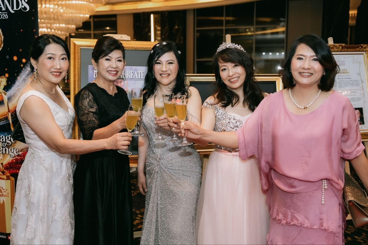 Left to right, a guest in a white lace gown, a key member of The BrandLaureate team in a black lace dress, DR. SHEKINAIH SIEW SIN YAP in her silver beaded gown with star pins, a guest in a pale pink gown and tiara and a guest in pink hold champagne glasses together and smile at the camera beside framed award portraits.