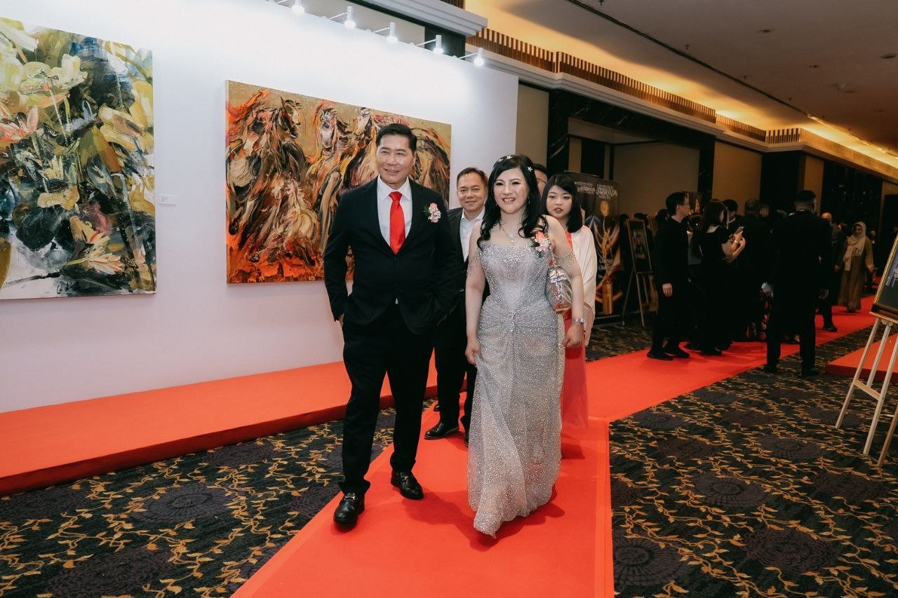 DR. J AARON K DAVID, in a dark suit and red tie, and DR. SHEKINAIH SIEW SIN YAP, in her silver beaded gown with a silver clutch, walk the red carpet along a corridor hung with large paintings, one of horses, with guests following behind and more guests gathered along the carpet.