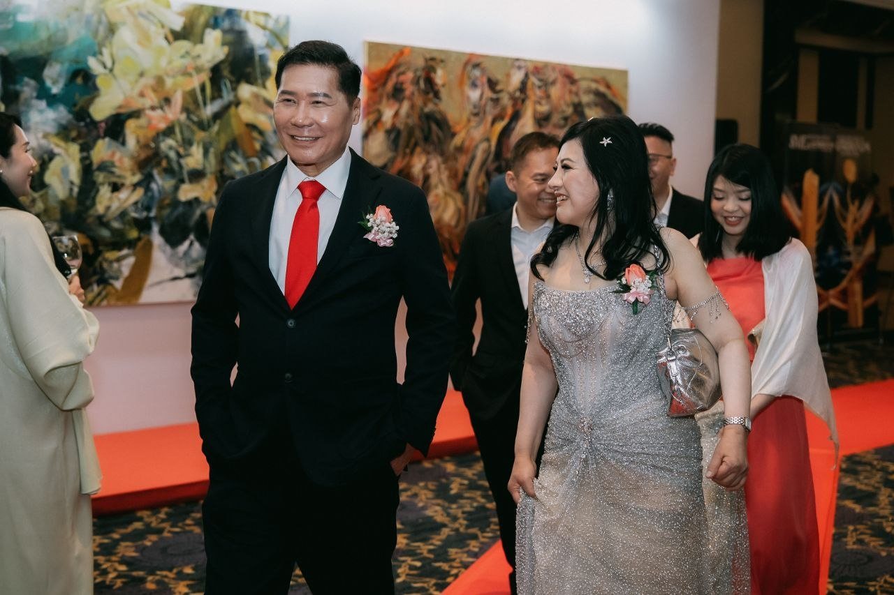 DR. J AARON K DAVID and DR. SHEKINAIH SIEW SIN YAP smile as they walk the red carpet along a corridor hung with large paintings, guests following