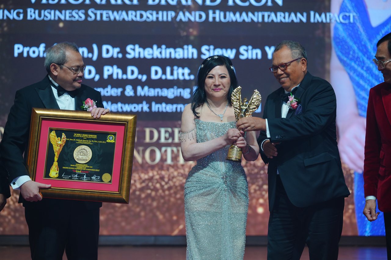 A presenter holds up the framed certificate while Tun Md Raus Sharif hands the gold trophy to DR. SHEKINAIH SIEW SIN YAP, beneath the screen with her name