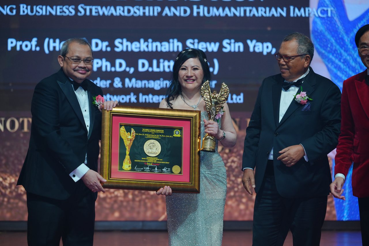DR. SHEKINAIH SIEW SIN YAP holds the gold trophy as a presenter beside her holds her framed certificate and Tun Md Raus Sharif stands at her other side, beneath the screen with her name