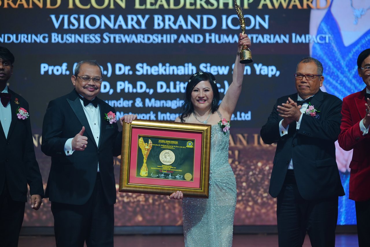 DR. SHEKINAIH SIEW SIN YAP raises the gold winged trophy and holds her framed certificate between a presenter giving a thumbs-up and Tun Md Raus Sharif applauding, with Prof. Dr. Edward Roy Krishnan standing at the left edge, beneath the screen reading Visionary Brand ICON, Enduring Business Stewardship and Humanitarian Impact, with her name