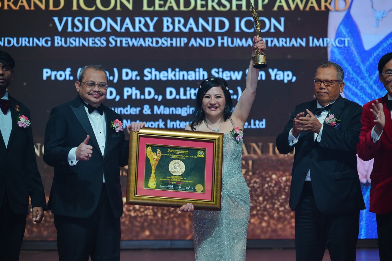 DR. SHEKINAIH SIEW SIN YAP raises the gold trophy and holds her framed certificate among the presenters, with Prof. Dr. Edward Roy Krishnan standing at the left edge and Tun Md Raus Sharif applauding beside her, beneath the screen reading Visionary Brand ICON
