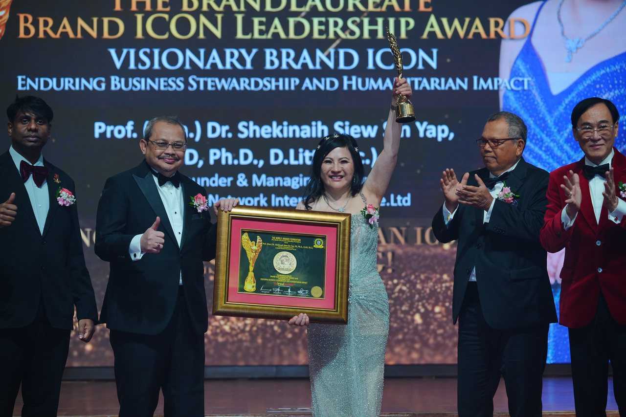 DR. SHEKINAIH SIEW SIN YAP lifts the trophy beneath the screen with her award title while Prof. Dr. Edward Roy Krishnan and a presenter on the left give a thumbs-up and Tun Md Raus Sharif and Prof. Dr. KKJohan applaud on the right