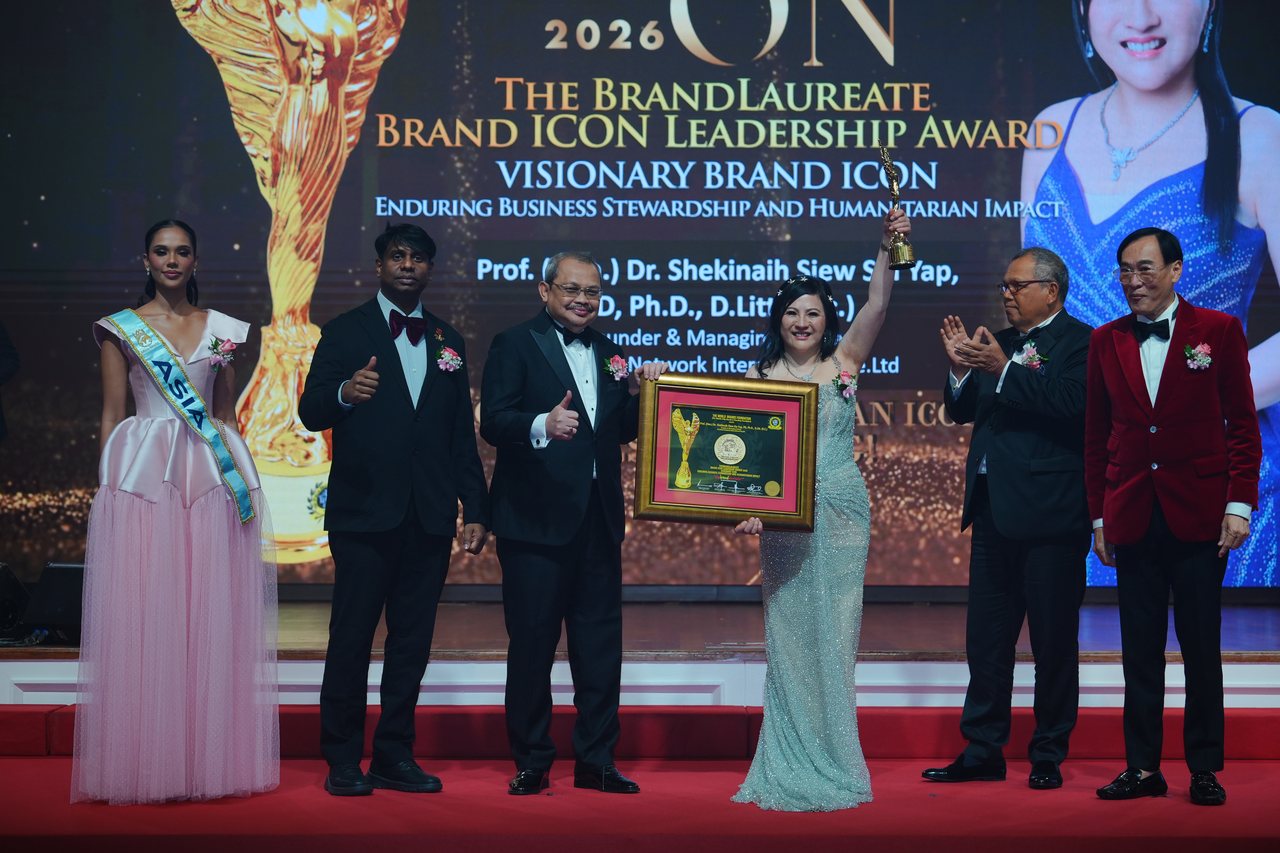 Left to right, Miss Universe Malaysia in a pale pink tulle gown and sash, Prof. Dr. Edward Roy Krishnan and a presenter giving a thumbs-up, the presenter sharing the framed certificate with DR. SHEKINAIH SIEW SIN YAP as she raises the trophy, Tun Md Raus Sharif applauding and Prof. Dr. KKJohan in his red velvet jacket stand on the red stage beneath the screen reading Visionary Brand ICON.