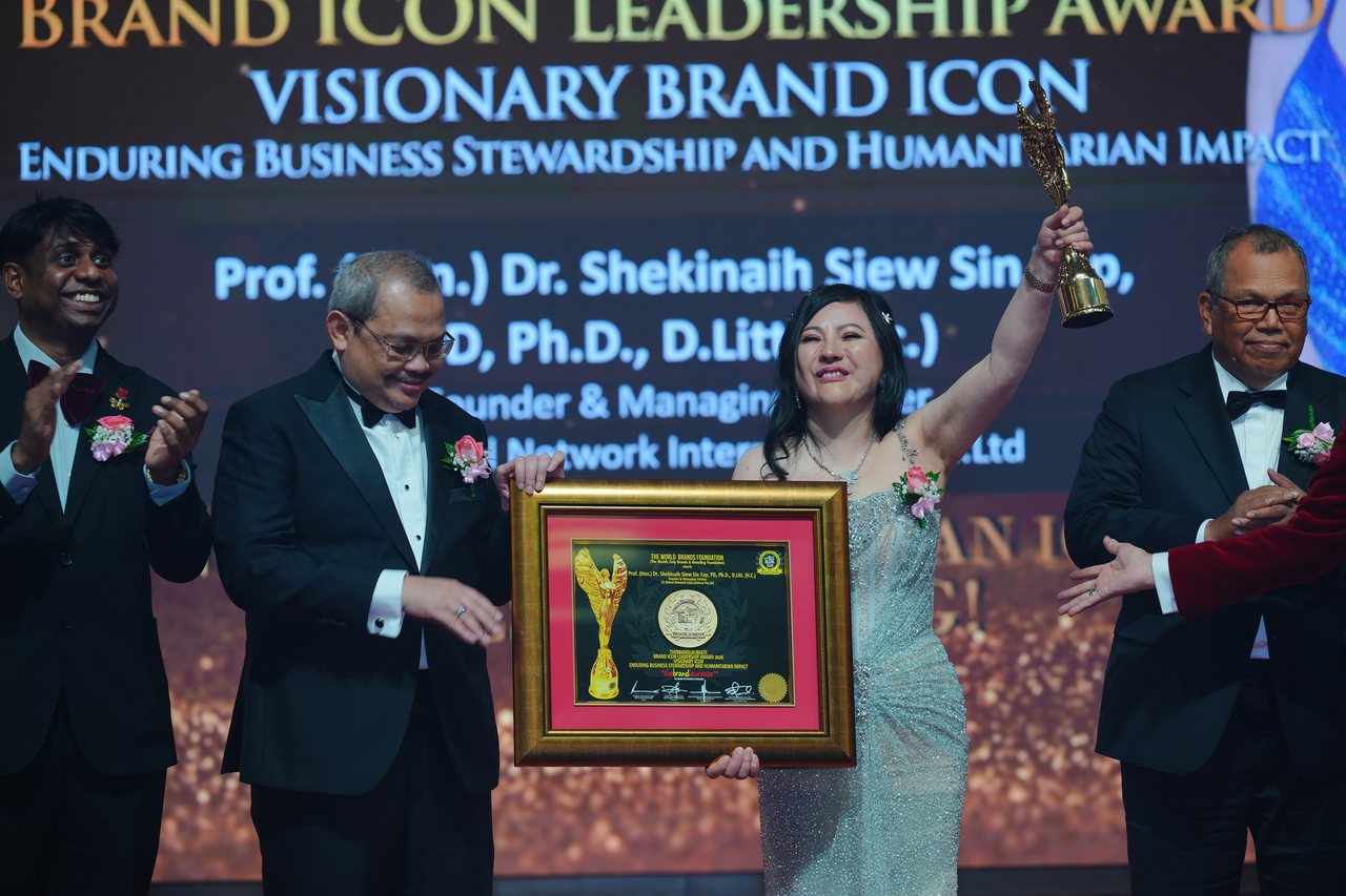 DR. SHEKINAIH SIEW SIN YAP raises the gold winged trophy with her framed certificate held in front of her, Prof. Dr. Edward Roy Krishnan applauding at the left and Tun Md Raus Sharif applauding at the right, beneath the screen reading Visionary Brand ICON, Enduring Business Stewardship and Humanitarian Impact