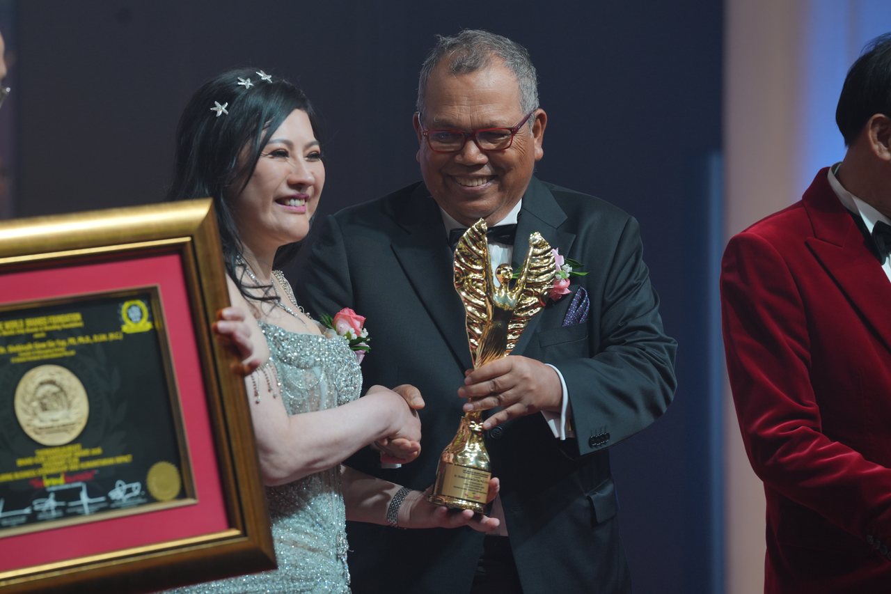 DR. SHEKINAIH SIEW SIN YAP smiles as Tun Md Raus Sharif shakes her hand and hands her the gold winged trophy, her framed certificate held up beside her