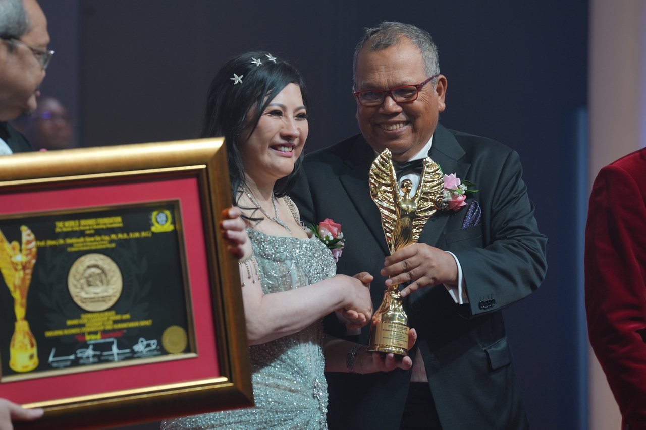 DR. SHEKINAIH SIEW SIN YAP smiles as she shakes hands with Tun Md Raus Sharif and takes the gold trophy, her framed certificate held beside her