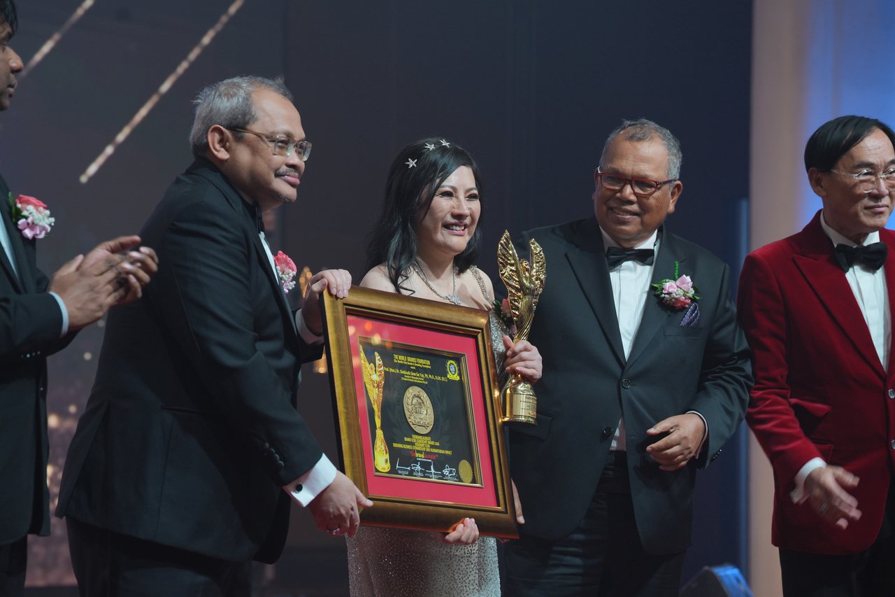 DR. SHEKINAIH SIEW SIN YAP smiles holding the gold trophy and her framed certificate, with a presenter holding the certificate beside her, Tun Md Raus Sharif standing at her side, Prof. Dr. Edward Roy Krishnan applauding at the left and Prof. Dr. KKJohan in a red velvet dinner jacket at the right.