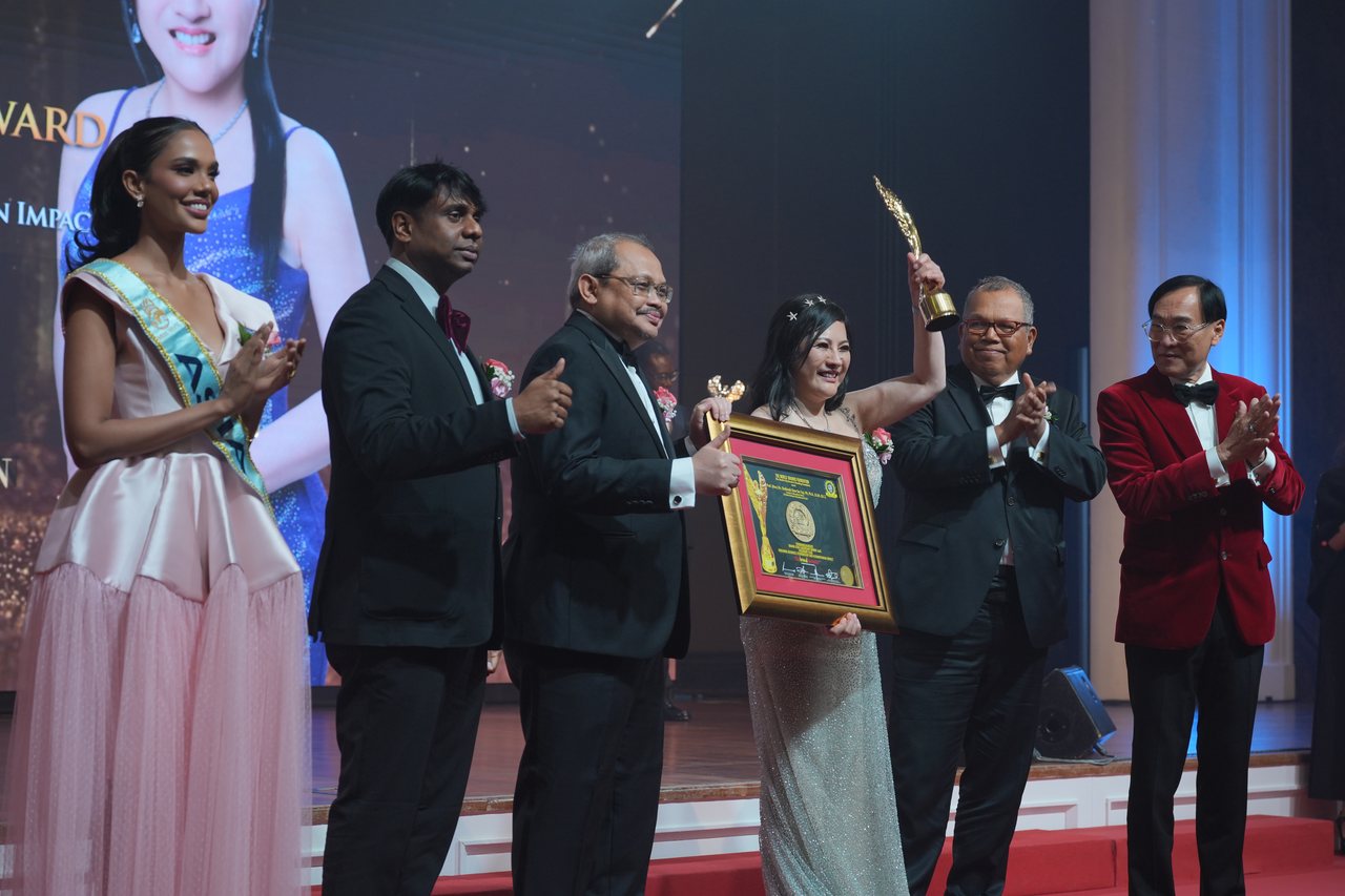 DR. SHEKINAIH SIEW SIN YAP raises the gold trophy on stage as Miss Universe Malaysia applauds, Prof. Dr. Edward Roy Krishnan and a presenter give a thumbs-up, and Tun Md Raus Sharif and Prof. Dr. KKJohan in a red velvet dinner jacket applaud beside her