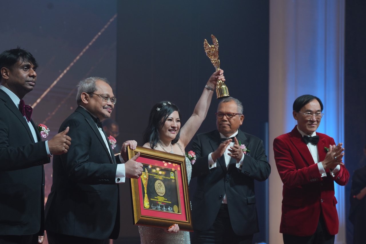 DR. SHEKINAIH SIEW SIN YAP lifts the gold trophy high as Prof. Dr. Edward Roy Krishnan and a presenter at the left give a thumbs-up, the presenter holding her framed certificate, and Tun Md Raus Sharif and Prof. Dr. KKJohan in a red velvet dinner jacket applaud at the right, seen from the side of the stage