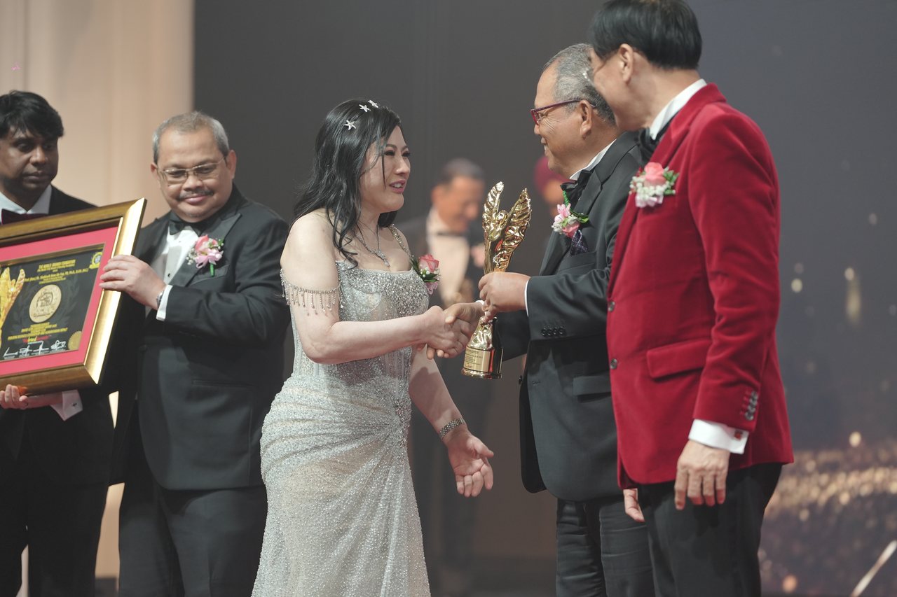 Seen from the side of the stage, DR. SHEKINAIH SIEW SIN YAP shakes hands with Tun Md Raus Sharif as he hands her the gold trophy, while a presenter holds her framed certificate, Prof. Dr. Edward Roy Krishnan stands behind it at the far left, and Prof. Dr. KKJohan in a red velvet dinner jacket stands in the foreground.