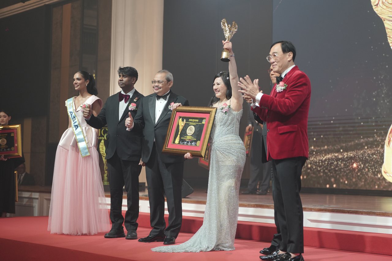Left to right, Miss Universe Malaysia in a pale pink tulle gown and sash, Prof. Dr. Edward Roy Krishnan in a maroon bow tie giving a thumbs-up, a presenter holding the framed certificate, DR. SHEKINAIH SIEW SIN YAP in her silver beaded gown and pink corsage raising the gold winged trophy overhead, and Prof. Dr. KKJohan in his red velvet dinner jacket applauding.