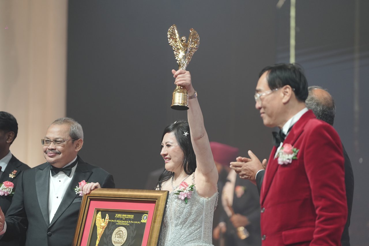 Close view of DR. SHEKINAIH SIEW SIN YAP smiling with the gold winged trophy raised high, Prof. Dr. Edward Roy Krishnan partly in view at the left edge, a presenter at left holding her framed certificate, Prof. Dr. KKJohan in his red velvet dinner jacket smiling at right and a presenter behind him applauding.