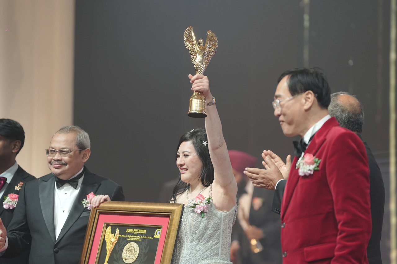 DR. SHEKINAIH SIEW SIN YAP raises the gold winged trophy high above her head as Prof. Dr. KKJohan, in a red velvet dinner jacket, stands beside her, a presenter behind him applauds, another presenter holds her framed certificate and Prof. Dr. Edward Roy Krishnan in his maroon bow tie stands at the left edge