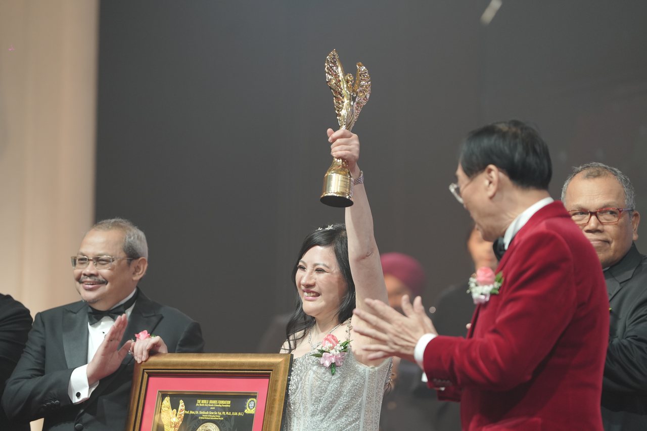 DR. SHEKINAIH SIEW SIN YAP smiles broadly with the gold winged trophy raised overhead, a presenter at left clapping beside her framed certificate, Prof. Dr. KKJohan in his red velvet dinner jacket applauding in the foreground and Tun Md Raus Sharif looking on at right.