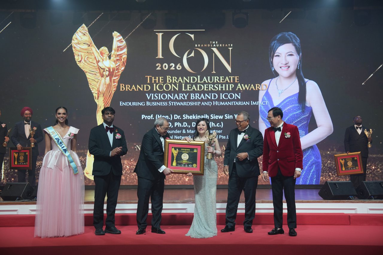 Left to right, Miss Universe Malaysia in a pale pink tulle gown and sash, Prof. Dr. Edward Roy Krishnan in a maroon bow tie applauding, a presenter handing the framed certificate to DR. SHEKINAIH SIEW SIN YAP as she holds the gold winged trophy and smiles, Tun Md Raus Sharif standing beside her, and Prof. Dr. KKJohan in his red velvet dinner jacket, beneath the screen with her portrait and the title Visionary Brand ICON.