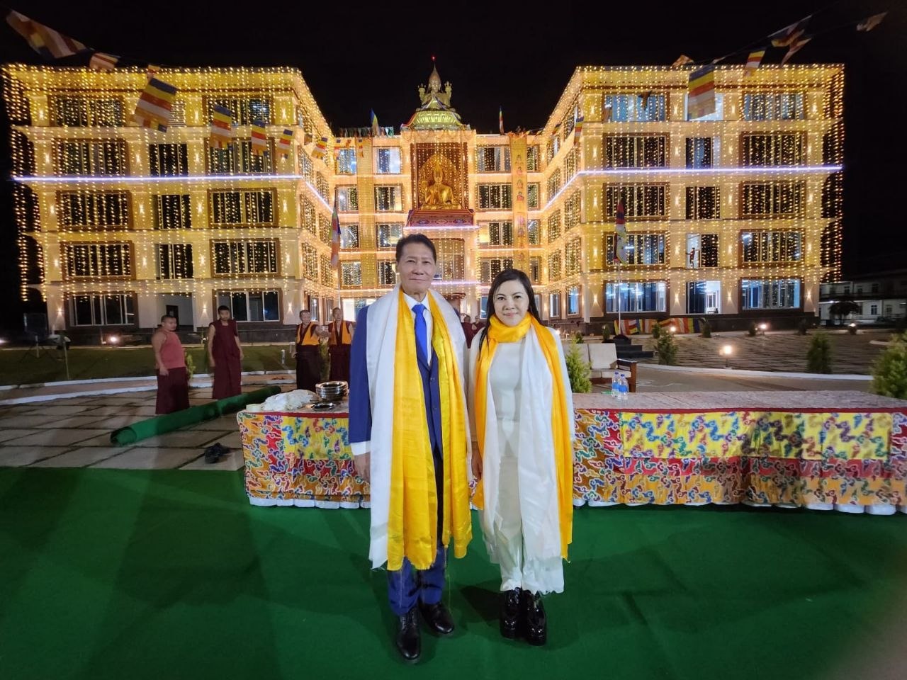 SHEKINAIH SIEW SIN YAP and J AARON K DAVID in front of the illuminated Ganden Monastery at night, wearing yellow khata from His Holiness the Dalai Lama