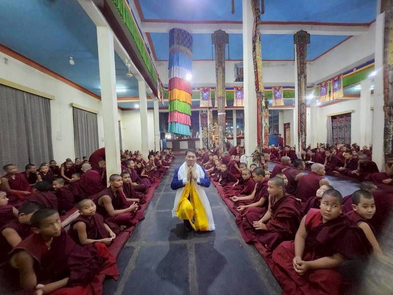 J AARON K DAVID kneeling in the central aisle of the temple, hands in prayer, surrounded by monks
