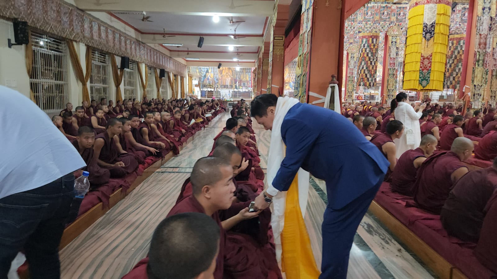 J AARON K DAVID handing financial offerings to seated monks in the prayer hall, Doeguling, Mundgod, January 2026
