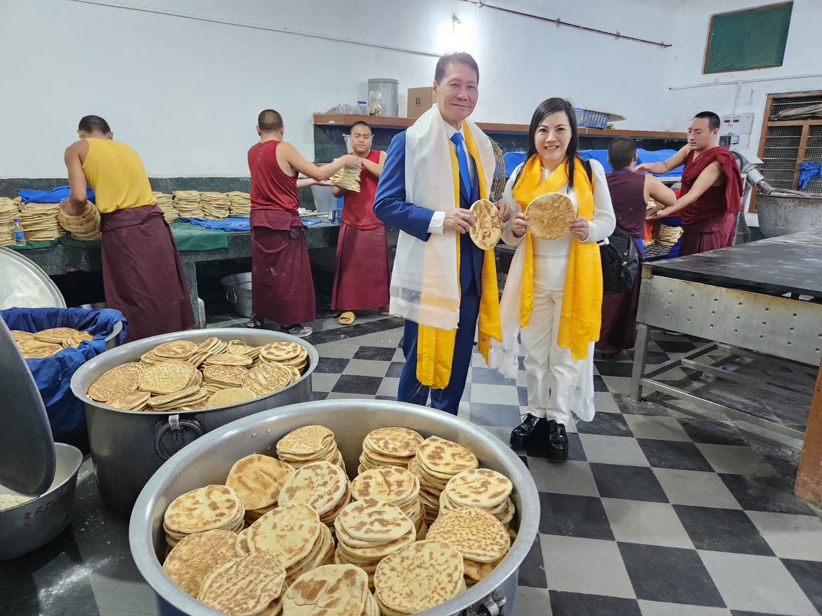 SHEKINAIH SIEW SIN YAP and J AARON K DAVID in the monastery kitchen holding fresh Tibetan flatbread, with monks baking large batches behind them, Doeguling, Mundgod, January 2026