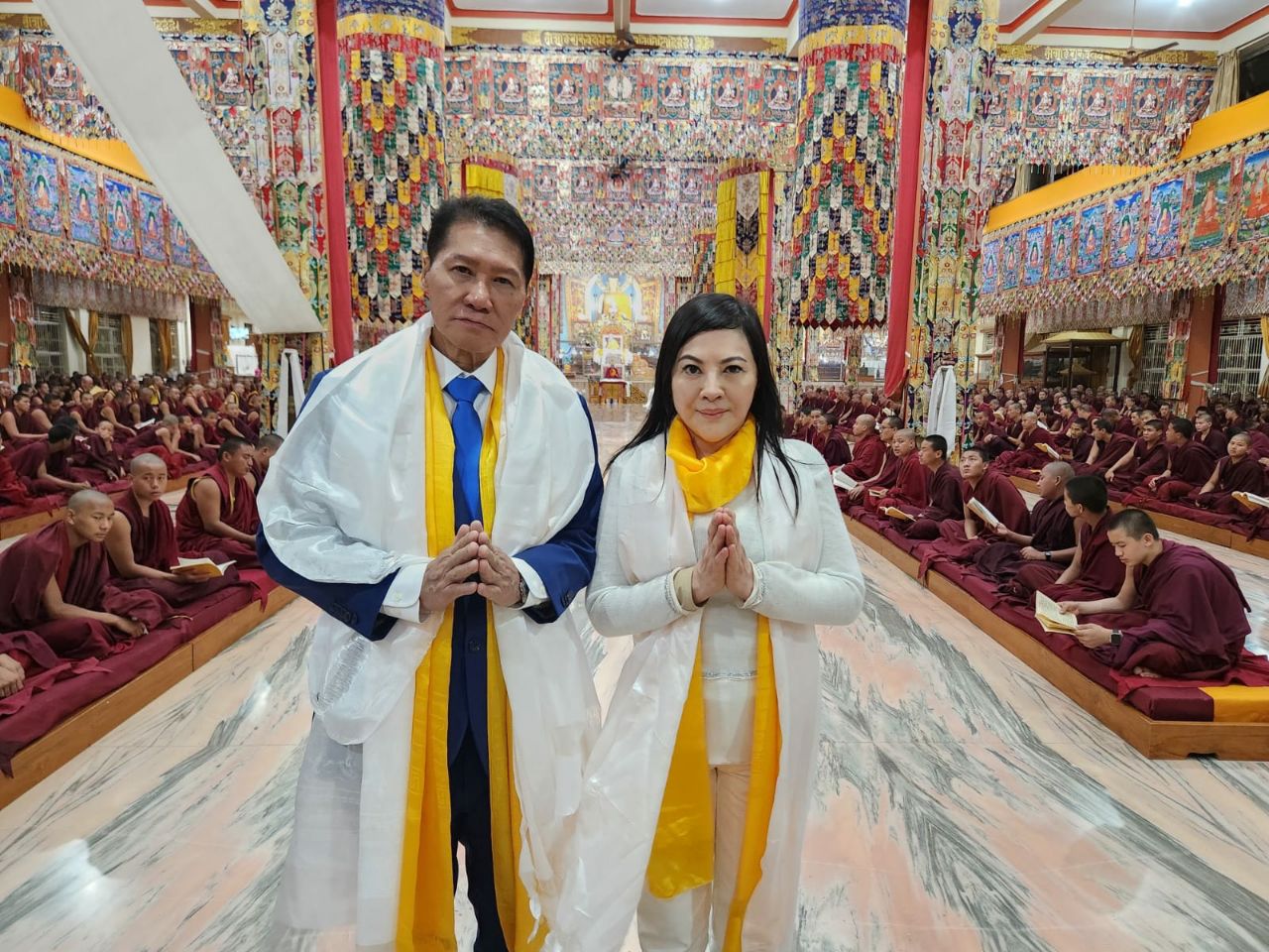 SHEKINAIH SIEW SIN YAP and J AARON K DAVID standing in añjali mudrā at the centre of the main thangka hall with hundreds of seated senior monks on either side, Doeguling Tibetan Settlement, Mundgod, January 2026
