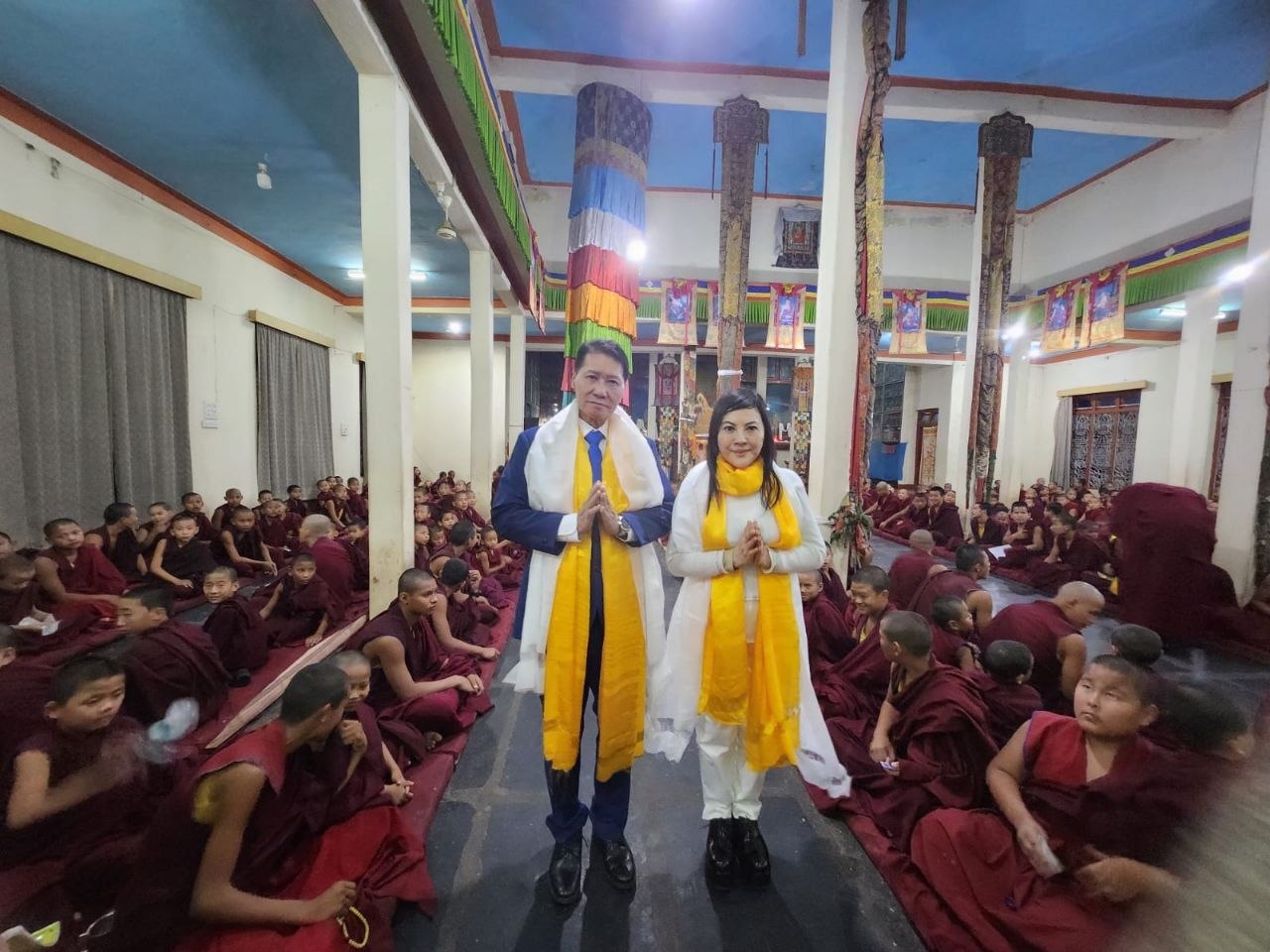 SHEKINAIH SIEW SIN YAP and J AARON K DAVID standing in añjali mudrā at the centre of the novice hall, surrounded by hundreds of young novice monks, Doeguling Tibetan Settlement, Mundgod, January 2026