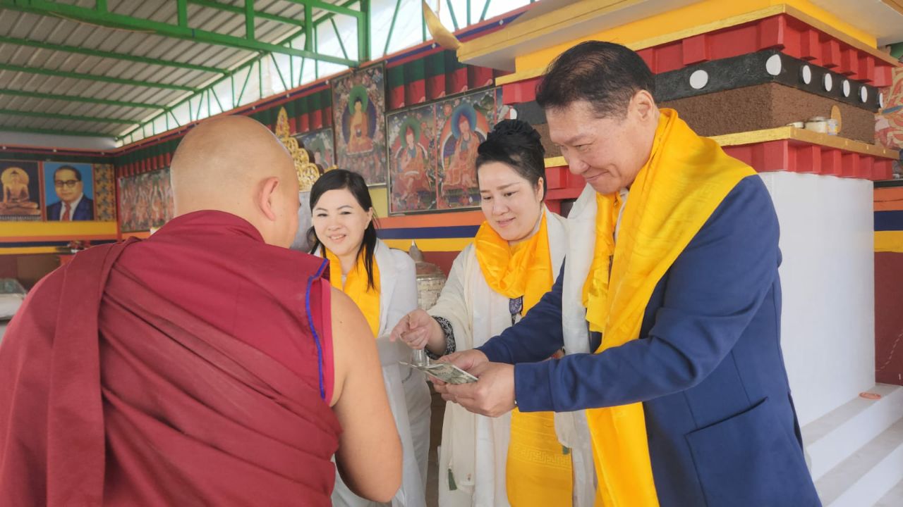 SHEKINAIH SIEW SIN YAP and J AARON K DAVID alongside a fellow delegate, presenting a financial offering into the hand of a senior monk at the outdoor stupa pavilion, Doeguling Tibetan Settlement, Mundgod, January 2026