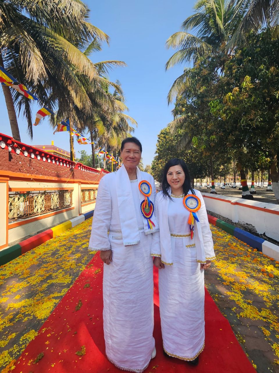 SHEKINAIH SIEW SIN YAP and J AARON K DAVID in white ceremonial robes with tri-colour Buddhist rosettes, standing on a red carpet strewn with yellow marigold petals beneath palm trees and Buddhist prayer flags at the Doeguling Tibetan Settlement entrance, Mundgod, January 2026
