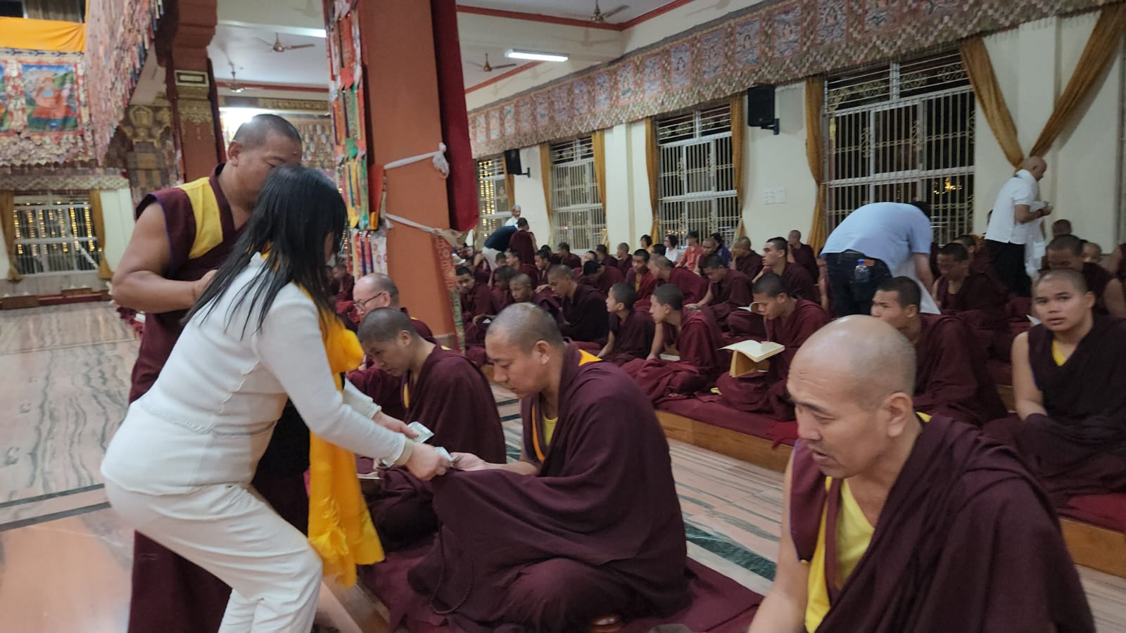 SHEKINAIH SIEW SIN YAP handing financial offerings to seated young monks in the prayer hall, Doeguling, Mundgod, January 2026