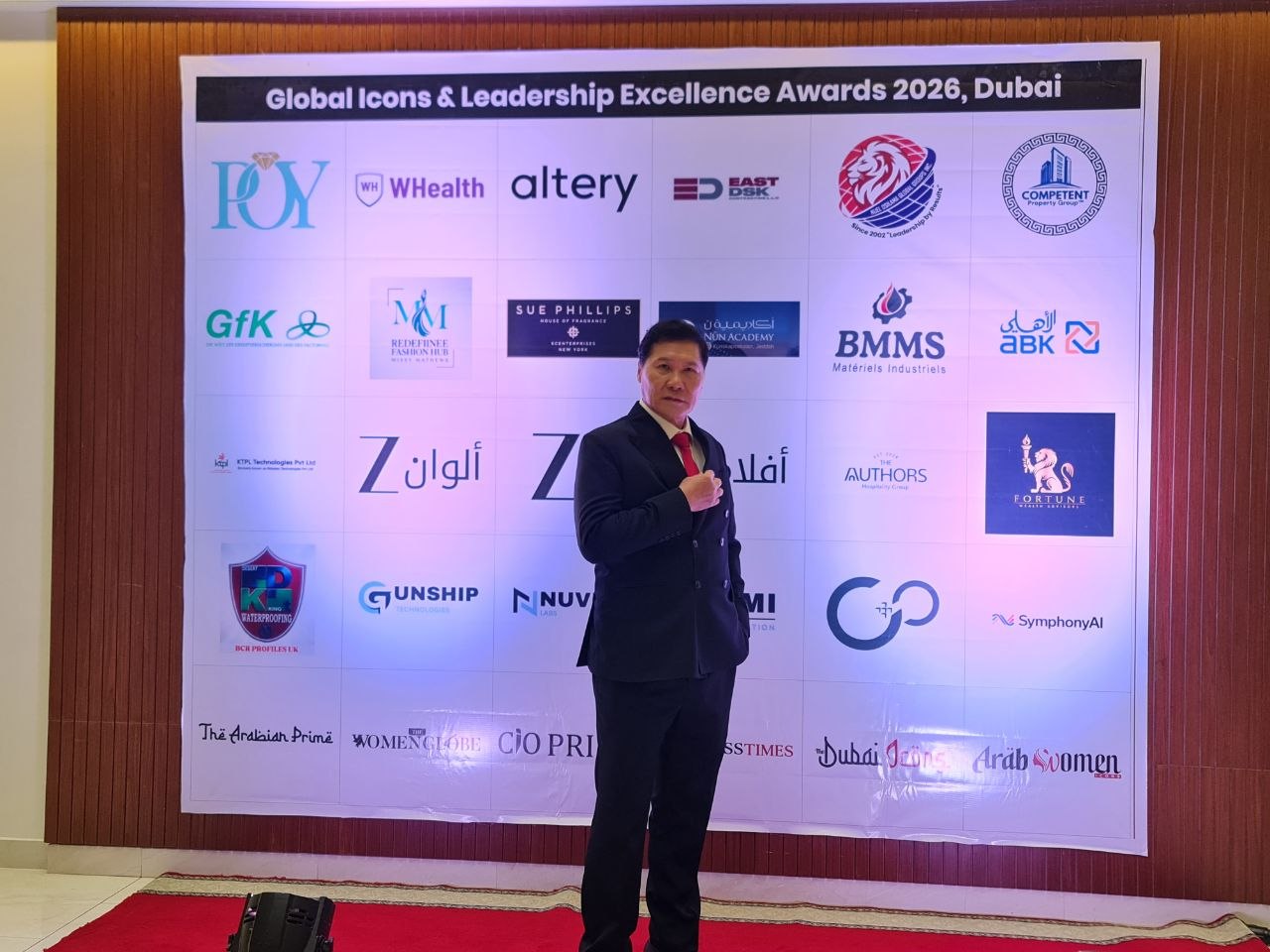 J AARON K DAVID in a dark suit and red tie before the Global Icons and Leadership Excellence Awards 2026 backdrop, Dubai