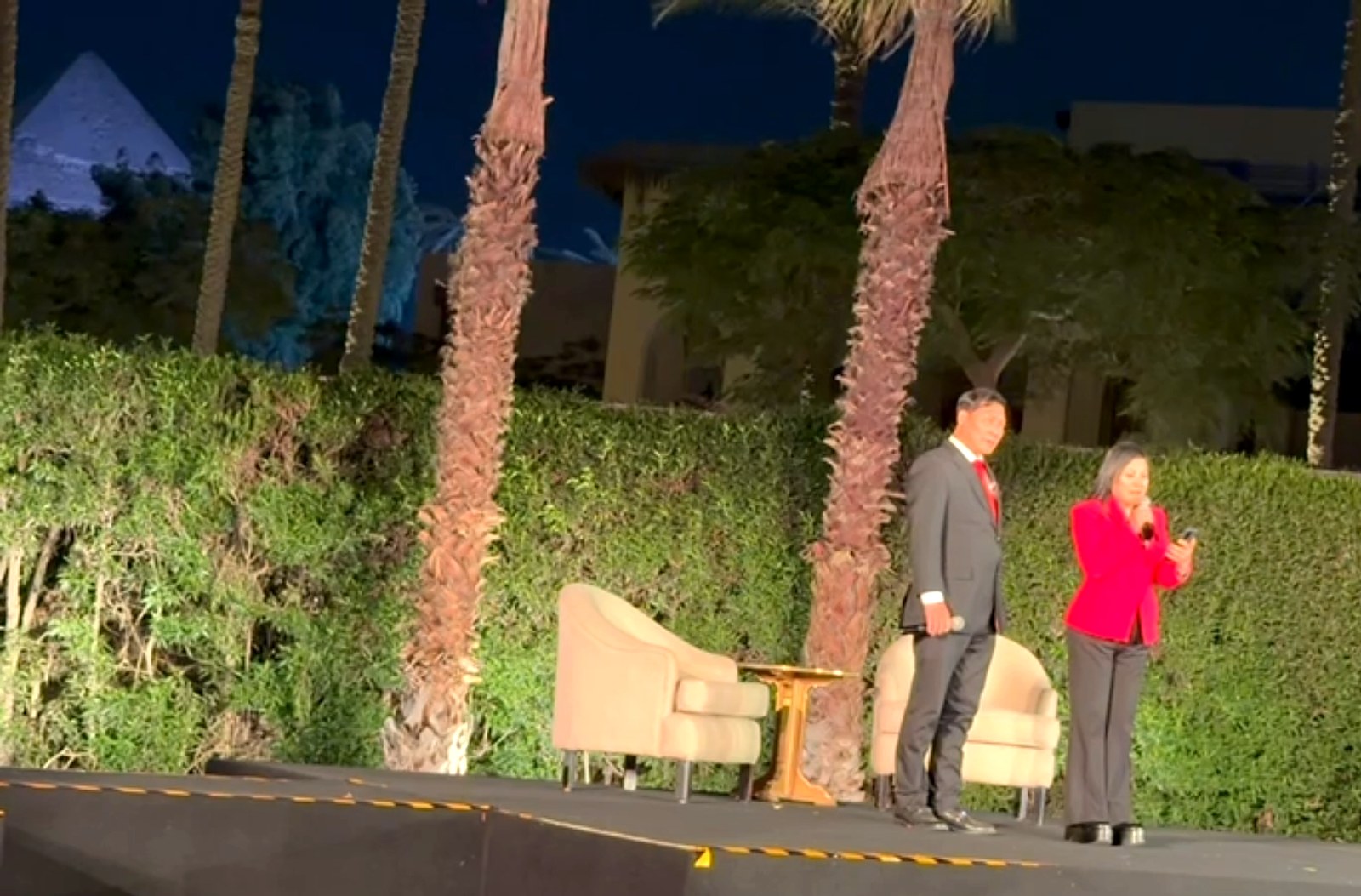 SHEKINAIH SIEW SIN YAP, in red, speaks on the open-air evening stage with J AARON K DAVID beside her; a lit pyramid rises beyond the palms at top left