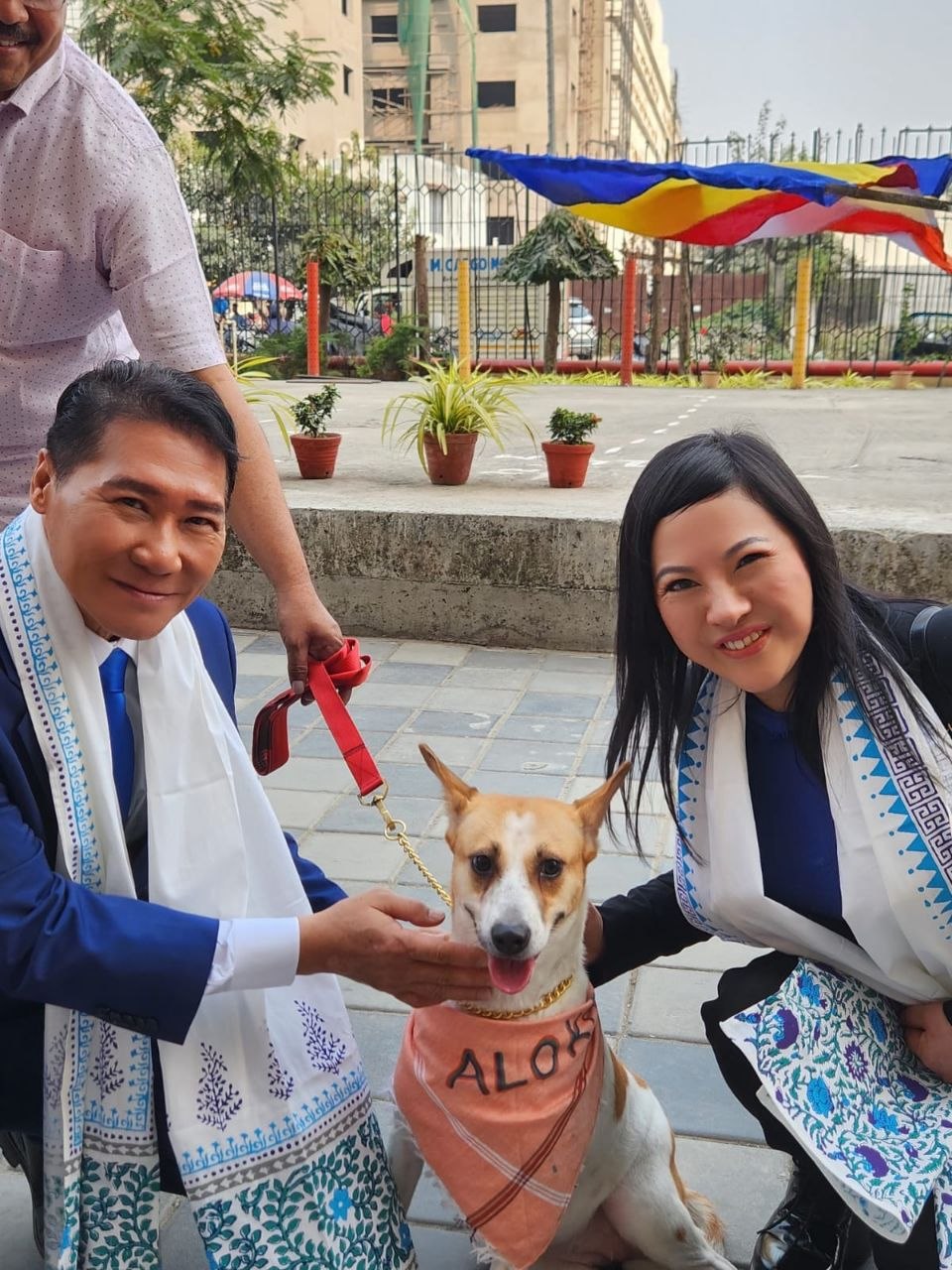 SHEKINAIH SIEW SIN YAP and J AARON K DAVID kneeling with Aloha, the rescued temple dog wearing the ALOHA bandana, beneath the Buddhist flag at the Walking-PEACE procession, Kolkata