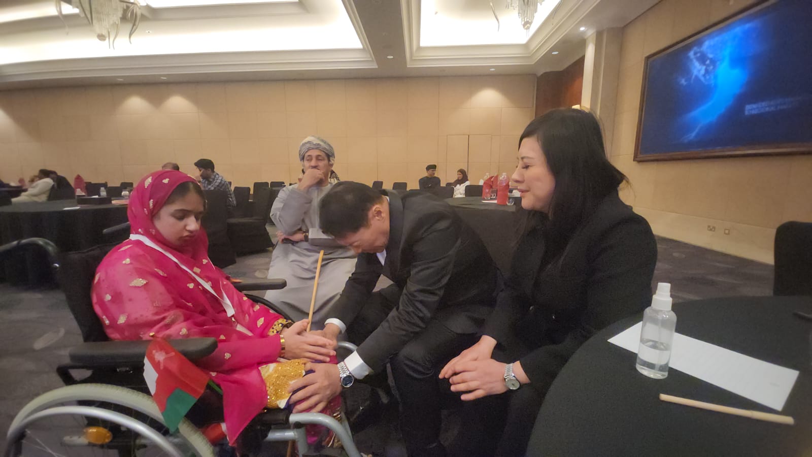 J AARON K DAVID leans in to pray with a young Omani girl in a wheelchair holding the Sultanate of Oman flag while SHEKINAIH SIEW SIN YAP sits beside in quiet presence