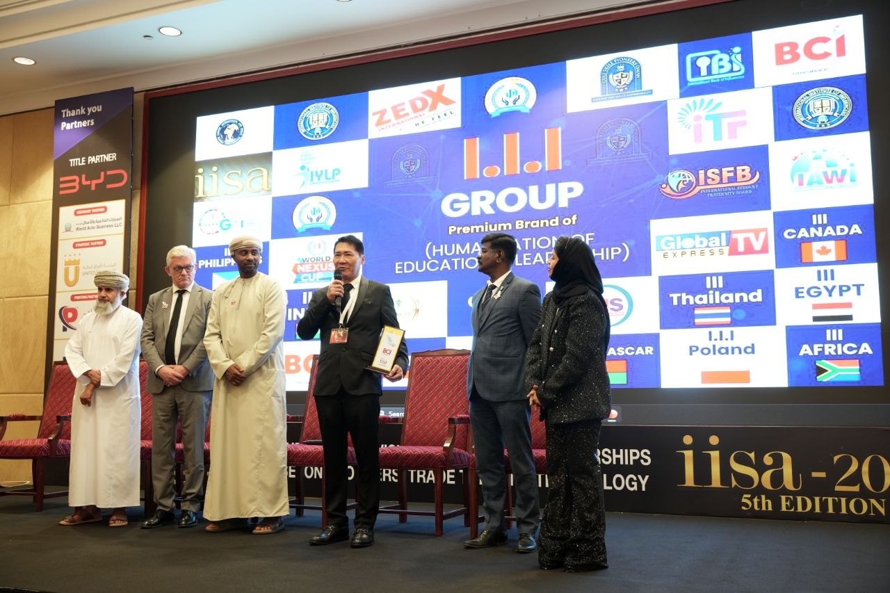 J AARON K DAVID speaking on the IISA 2026 stage with international delegate panel — Omani host, European partner, Indian delegate, and a Thai representative